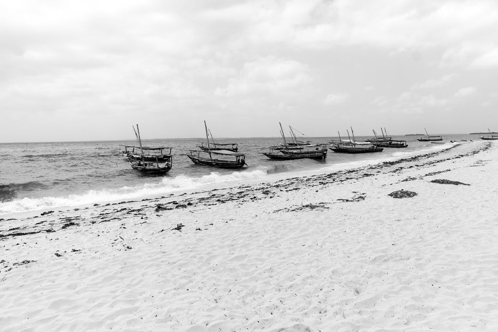 The FN Prints "Tourist Boats At Nakupenda Sandbank" captures the coastal elegance of a black and white scene featuring several small wooden boats anchored near the shore. The sandy beach, sprinkled with seaweed, lies under a calm ocean and a cloudy sky.