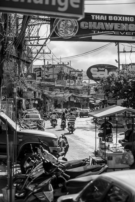 A bustling street scene in FN Prints' "A Chaweng Street: Koh Samui" showcases Thailand's Chaweng Street filled with numerous motorcycles, vendors, and a plethora of signs, including one advertising Thai boxing at Chaweng Stadium. Overhead wires crisscross the lively area teeming with activity amid densely packed buildings. The black and white photography beautifully captures the vibrant atmosphere.