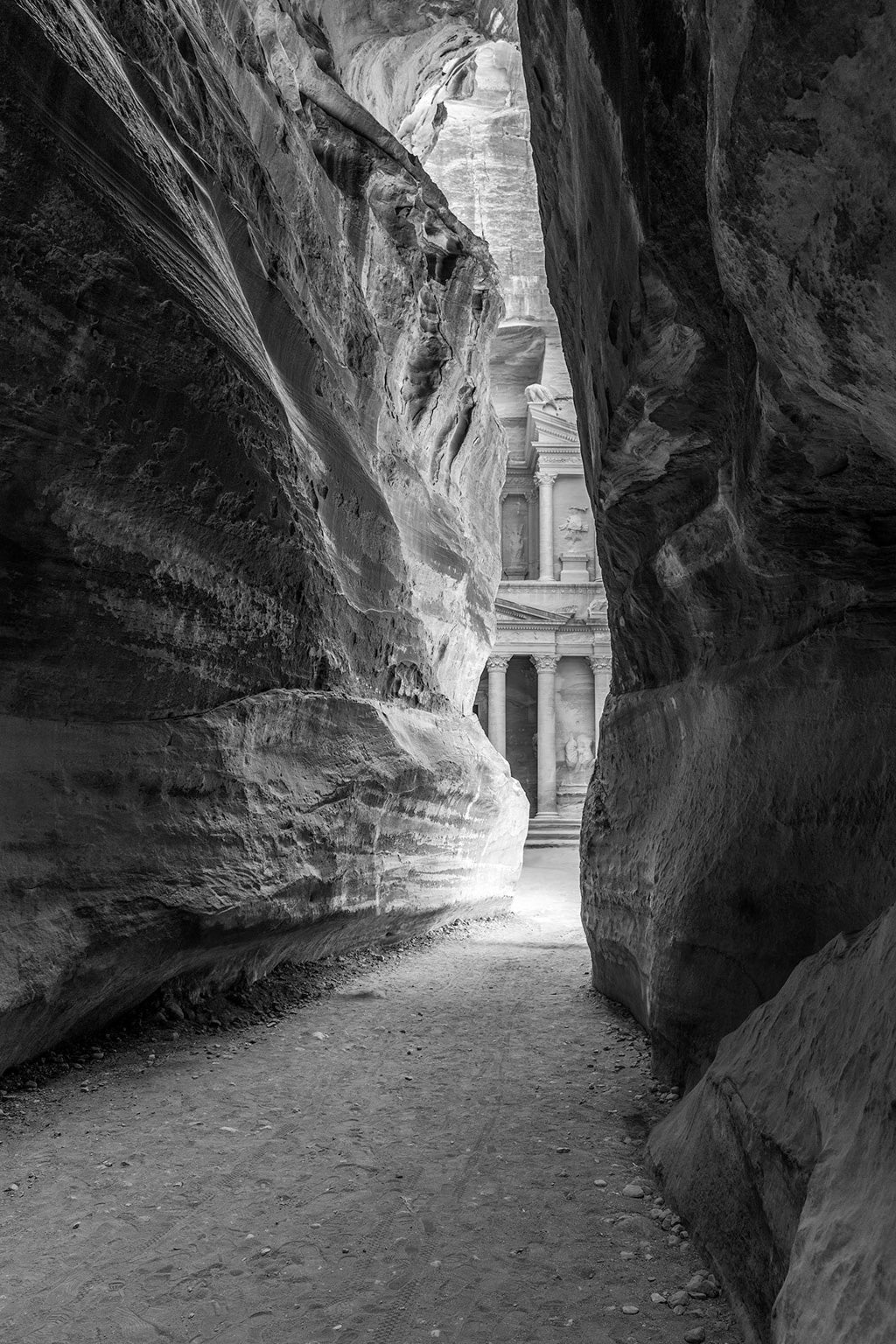 The product "A Peak of Al-Khazneh - The Treasury Petra Black & White" by FN Prints features a black and white photograph showcasing a narrow canyon walkway that leads to the famous Al-Khazneh (The Treasury) in Petra, Jordan. Tall, weathered rock walls frame the path, and sunlight illuminates the intricately carved façade of the ancient structure in the background.