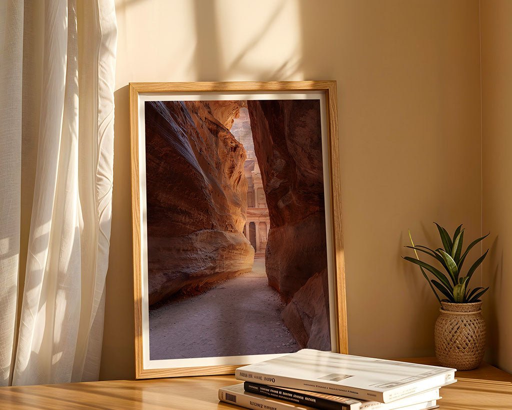 A framed photograph titled "A Peak Of Al-Khazneh - The Treasury Petra" by FN Prints, capturing a narrow sandstone canyon with streaming light, is placed on a wooden table. Next to the frame are stacked books and a small potted plant. Sunlight filters through a sheer curtain, casting soft shadows on the wall.