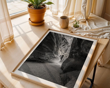 A framed black-and-white photograph titled "A Peak Of Al-Khazneh - The Treasury Petra" by FN Prints is displayed on a wooden table. The table also features a potted plant, a small cup, and some fabric near a sunlit window with sheer curtains.