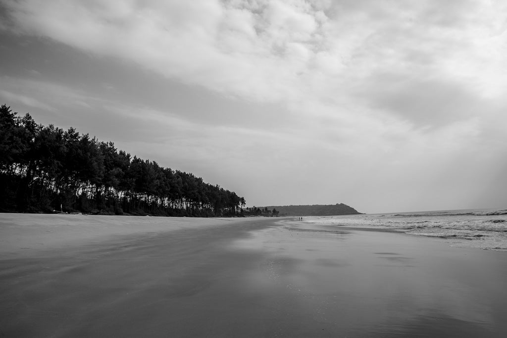 A Walk On The Beach" by FN Prints captures a tranquil black and white scene of an empty Goan coastal retreat, where gentle waves kiss the shoreline. A dense line of trees stretches along the left edge, fading into the horizon under an overcast sky. Beams of light pierce through the clouds, casting reflections on the damp sand.