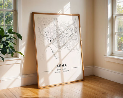 In a sunlit room, an Abha City Map by FN Prints leans against a wall on wooden flooring, shadows play over the museum-quality paper. The monochrome map intricately shows city streets with geographical coordinates subtly printed below.