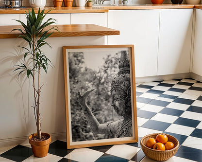 An FN Prints framed photograph titled "Abhaya Mudra," depicting a Thai Buddha statue in black and white, is propped against a kitchen counter. The kitchen itself showcases checkered black and white flooring, complemented by white cabinets and a wooden counter. A potted plant alongside a bowl of oranges on the floor contributes to the space's spiritual symbolism.