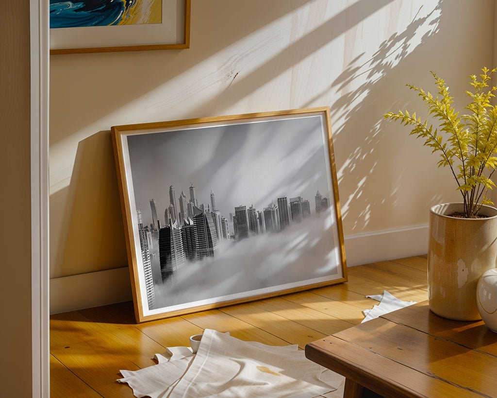 In a sunlit room, FN Prints' "Above The Clouds," a framed piece of black-and-white photography capturing Dubai's high-rises, leans against a wall. Nearby, a wooden table and a potted plant with yellow flowers cast intricate shadows on the wooden floor.