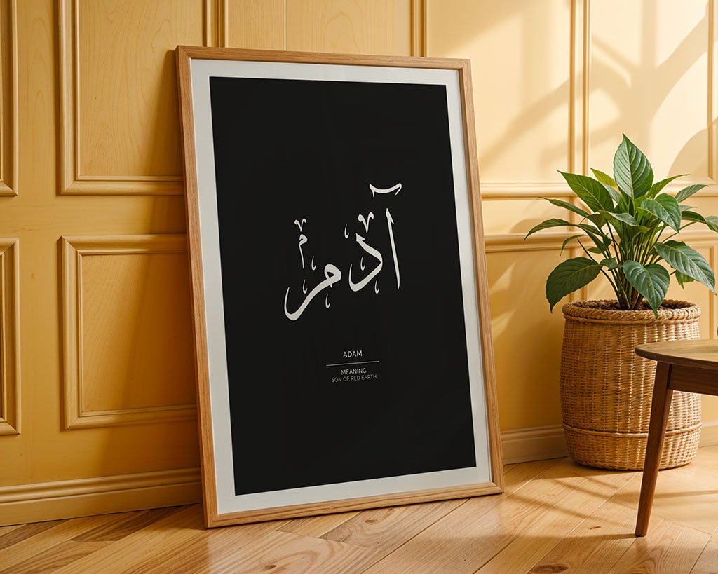 A framed Adam - آدم wall art poster by FN Prints, featuring Arabic calligraphy and the meaning of "Adam," leans against a yellow paneled wall beside a potted plant on a wooden floor. Part of the Arabic Names Collection.