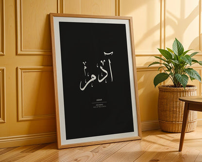 A framed Adam - آدم wall art poster by FN Prints, featuring Arabic calligraphy and the meaning of "Adam," leans against a yellow paneled wall beside a potted plant on a wooden floor. Part of the Arabic Names Collection.