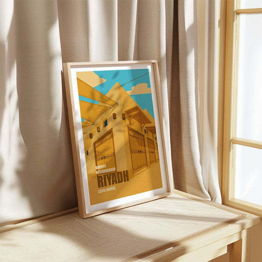 Framed artwork of a street scene with 'Riyadh' text, placed on a wooden surface with curtains in the background.