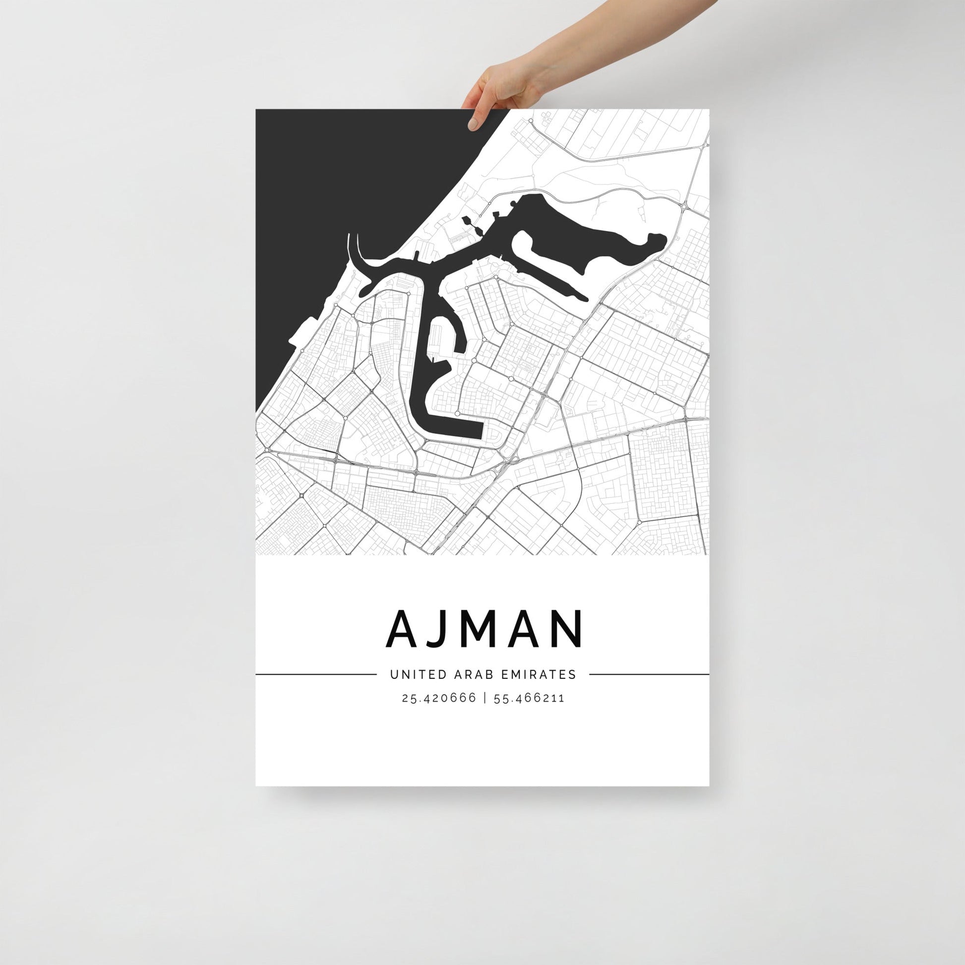 A person is holding the FN Prints Ajman Map, a black and white wall art print showcasing detailed streets and waterways of Ajman, United Arab Emirates. The design features geographic coordinates 25.420066 | 55.466211 beneath this artistic representation of one of UAE’s hidden gems.