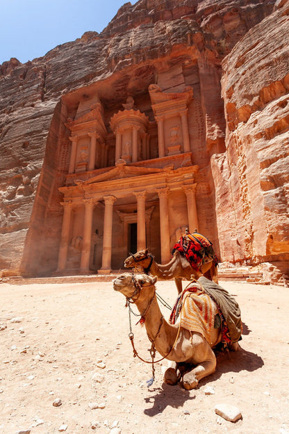 Two camels rest in front of Al-Khazneh - The Treasury, an ancient rock-carved temple and architectural marvel in Jordan. The elaborate façade features columns and intricate carvings against a backdrop of towering, rugged cliffs. The camels are adorned with colorful, patterned blankets and saddles. This stunning scene is beautifully captured by FN Prints in their product, "Al-Khazneh - The Treasury - Petra: Part I.