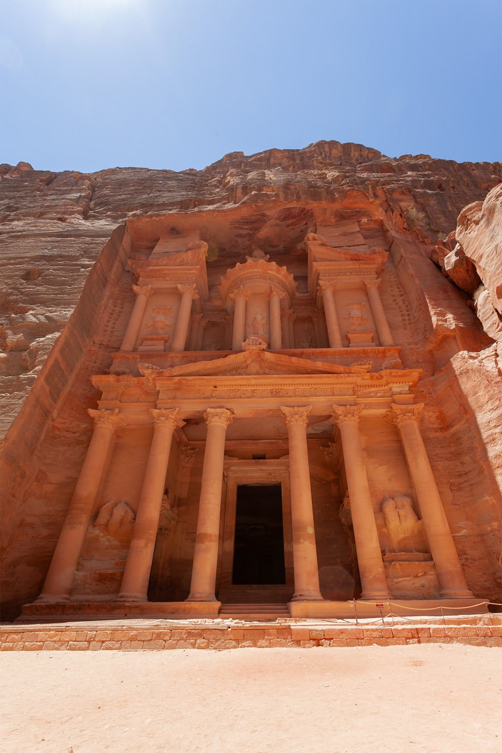 The FN Prints product "Al-Khazneh - The Treasury - Petra: Part II" showcases a vibrant color photograph of the ancient city of Petra in Jordan, specifically highlighting Al-Khazneh (The Treasury). The image captures the intricate façade carved into red sandstone cliffs, adorned with elaborate architectural elements such as columns and detailed carvings, all set against a clear, blue sky.