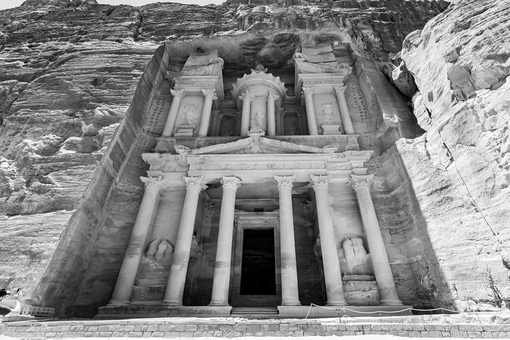 Black and white photograph from FN Prints titled "Al-Khazneh - The Treasury - Petra: Part III," showcasing the ancient wonder carved into the sandstone rock face at Petra, Jordan. This masterpiece features intricate columns, sculptures, and carvings framed by towering cliff walls, with shadows enhancing its majestic appearance.