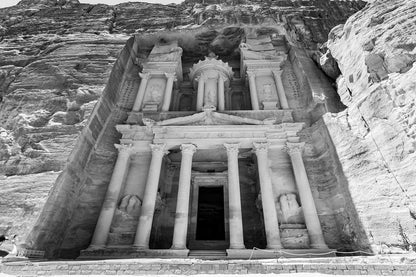 Black and white photograph from FN Prints titled "Al-Khazneh - The Treasury - Petra: Part III," showcasing the ancient wonder carved into the sandstone rock face at Petra, Jordan. This masterpiece features intricate columns, sculptures, and carvings framed by towering cliff walls, with shadows enhancing its majestic appearance.