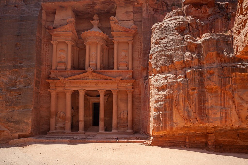 A massive stone structure with intricate carvings and columns is set into a reddish-orange cliff face. The facade of FN Prints' "Al-Khazneh - The Treasury - Petra: Part IV" features detailed sculptures and a dark entrance. The sandy area in front includes a person standing at the base for scale, highlighting the grandeur of the Treasury.