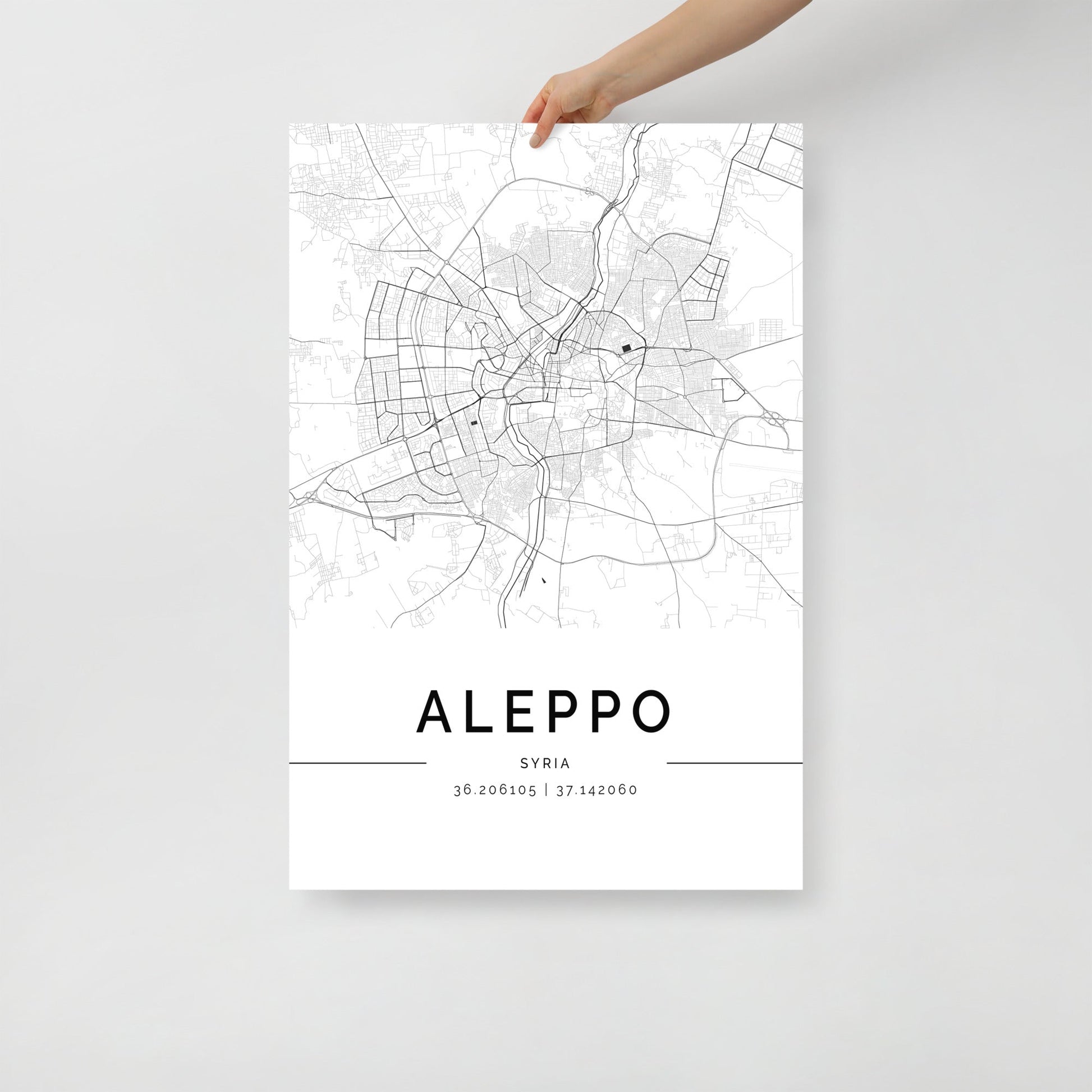 A hand holds an FN Prints' Aleppo Map poster, showcasing intricate street details along with geographic coordinates at the bottom. This elegant wall art print features a minimalist design with a plain, light background, making it perfect for any decor.