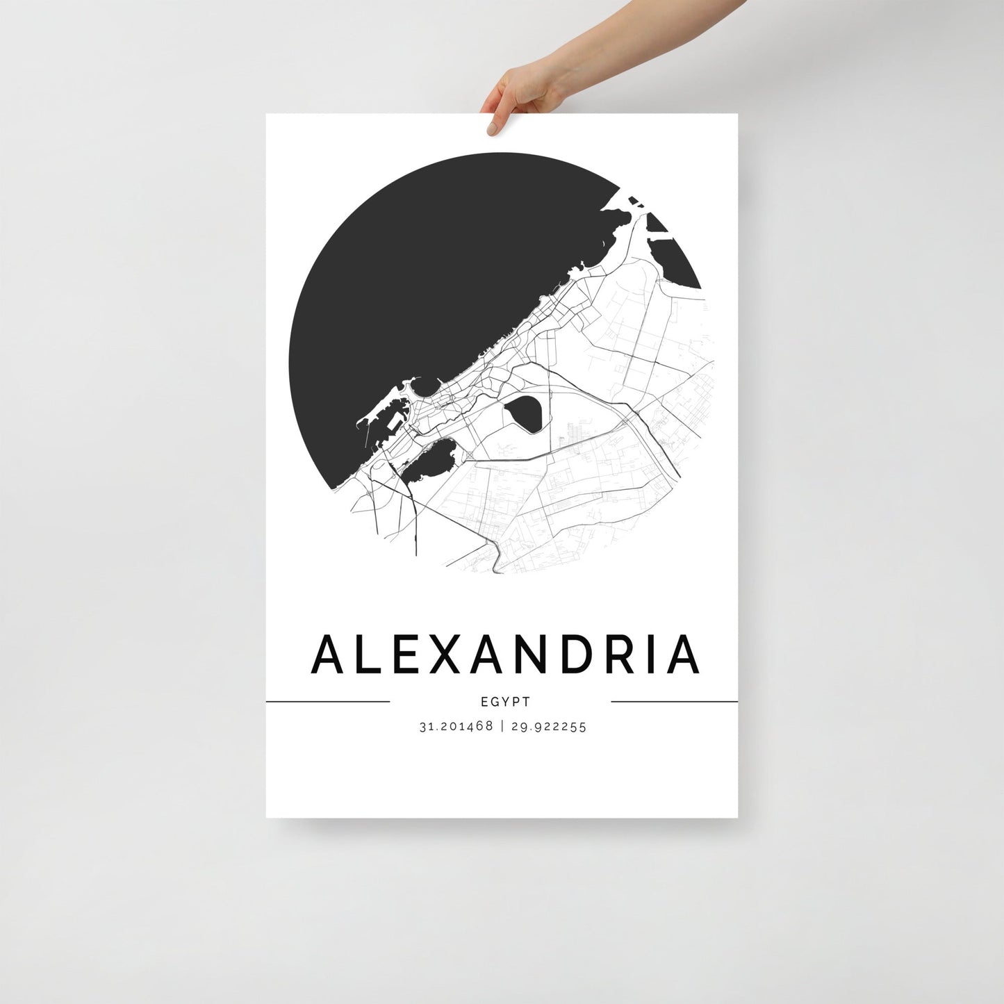 A person holds a minimalist Alexandria Map poster by FN Prints, showcasing a black and white depiction of the historic city in Egypt with the geographic coordinates 31.201468, 29.922255 displayed.