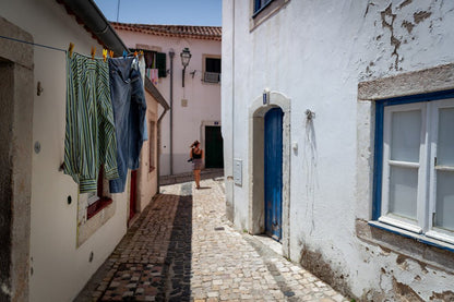 A narrow cobblestone street in a quaint village with whitewashed buildings, blue doors, and clothes drying on a line exudes serene simplicity. A person stands in the distance, looking at something on the side of a building, capturing the cultural authenticity reminiscent of Alfama. The scene is bright with natural daylight—a perfect depiction found in the Alfama Explorer by FN Prints.