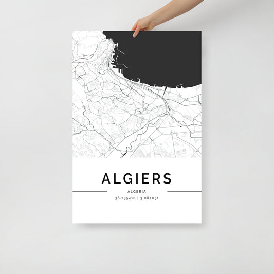 A person holds the FN Prints Algiers Map wall art print. Featuring a minimalist black and white design, it highlights detailed street lines with the sea depicted in dark shading. Below the city's name are the precise coordinates: 36.75341° N, 3.084051° E.