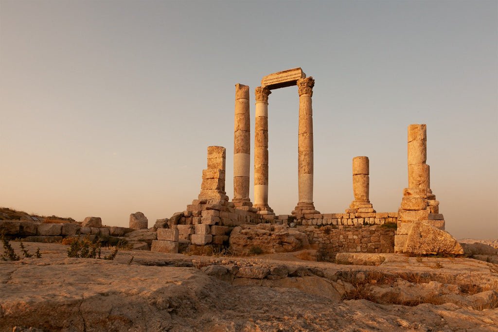 FN Prints' depiction of the Amman Citadel features ancient stone columns standing among Roman ruins at sunset. The pillars vary in height, with a clear, golden sky casting long shadows on the rocky ground. This historical site is beautifully captured, with some structures partially intact.