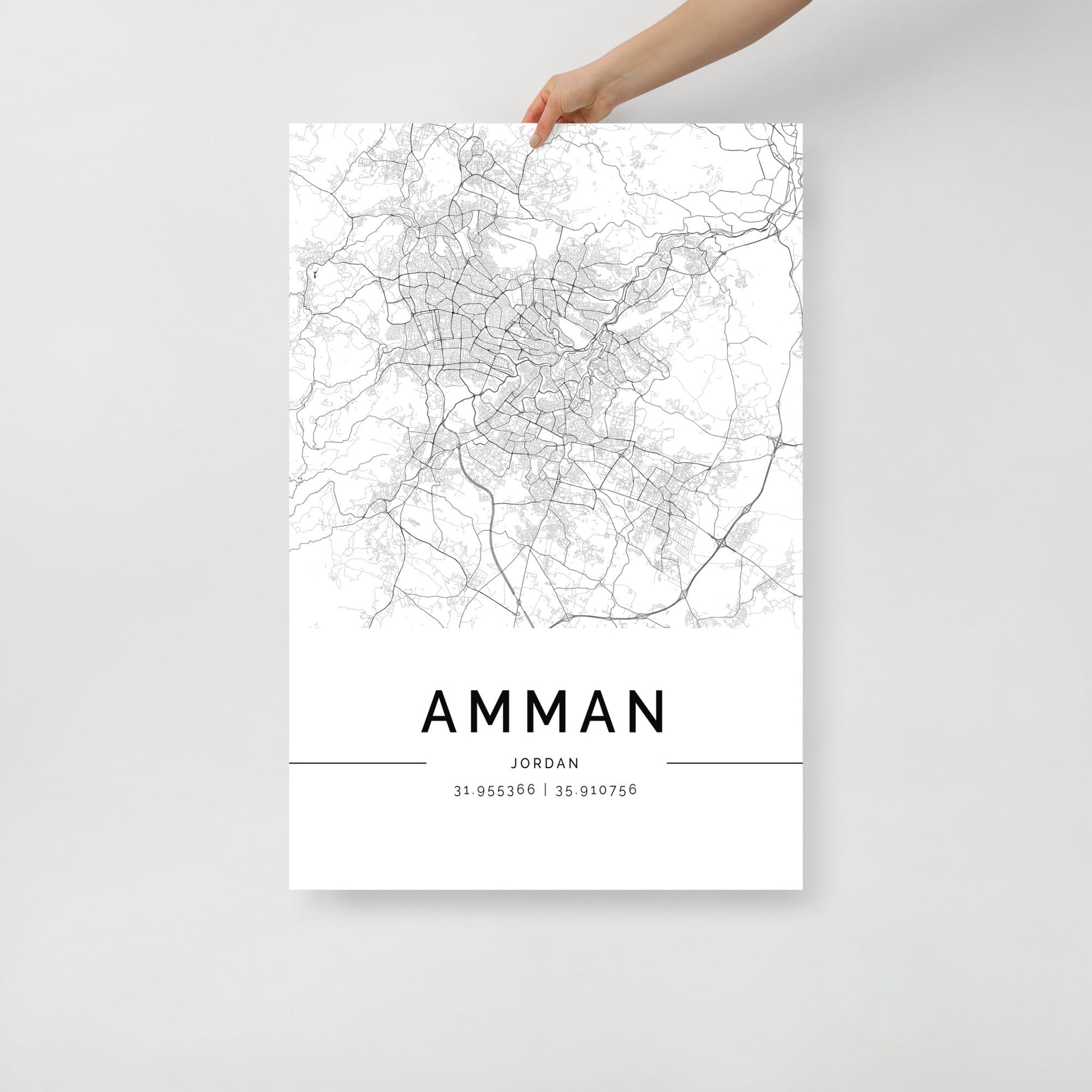 A hand showcases an elegant piece of wall art featuring FN Prints' Amman City Map, a detailed black and white depiction of the city with geographic coordinates 31.95356°N, 35.91076°E beneath the name, capturing the charm of Middle Eastern architecture.