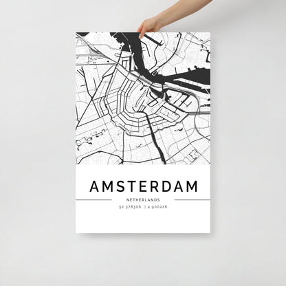 A hand displays the Amsterdam Map wall art from FN Prints, featuring a black and white poster that highlights the detailed street and canal designs of Amsterdam. The map is labeled with "AMSTERDAM, NETHERLANDS" followed by coordinates 52.37936 | 4.900226 at its base.