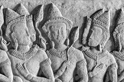 A detailed stone relief from FN Prints, titled "Ancient Reliefs: Angkor Wat," depicts a group of human figures adorned with intricate headgear and jewelry, reminiscent of historical art. They hold staffs and appear to be engaged in a dialogue or procession, with craftsmanship that highlights the delicate features and patterns on their attire.