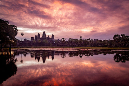 A stunning sunrise over Angkor Wat in Cambodia, reflected in a calm lake, creates a serene and majestic scene. The sky is painted with vibrant hues of pink, purple, and orange. This Angkor Wat Sunrise by FN Prints captures the silhouette of the temple surrounded by palm trees perfectly.