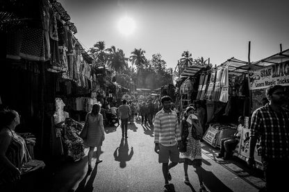 The black and white photograph, titled "Anjuna Market Hustle" by FN Prints, encapsulates the vibrant energy of vendors selling clothes and various items on either side. Shoppers meander through Anjuna Market beneath a radiant sun, while palm trees sway in the background, heightening the lively ambiance of this iconic Goa locale.