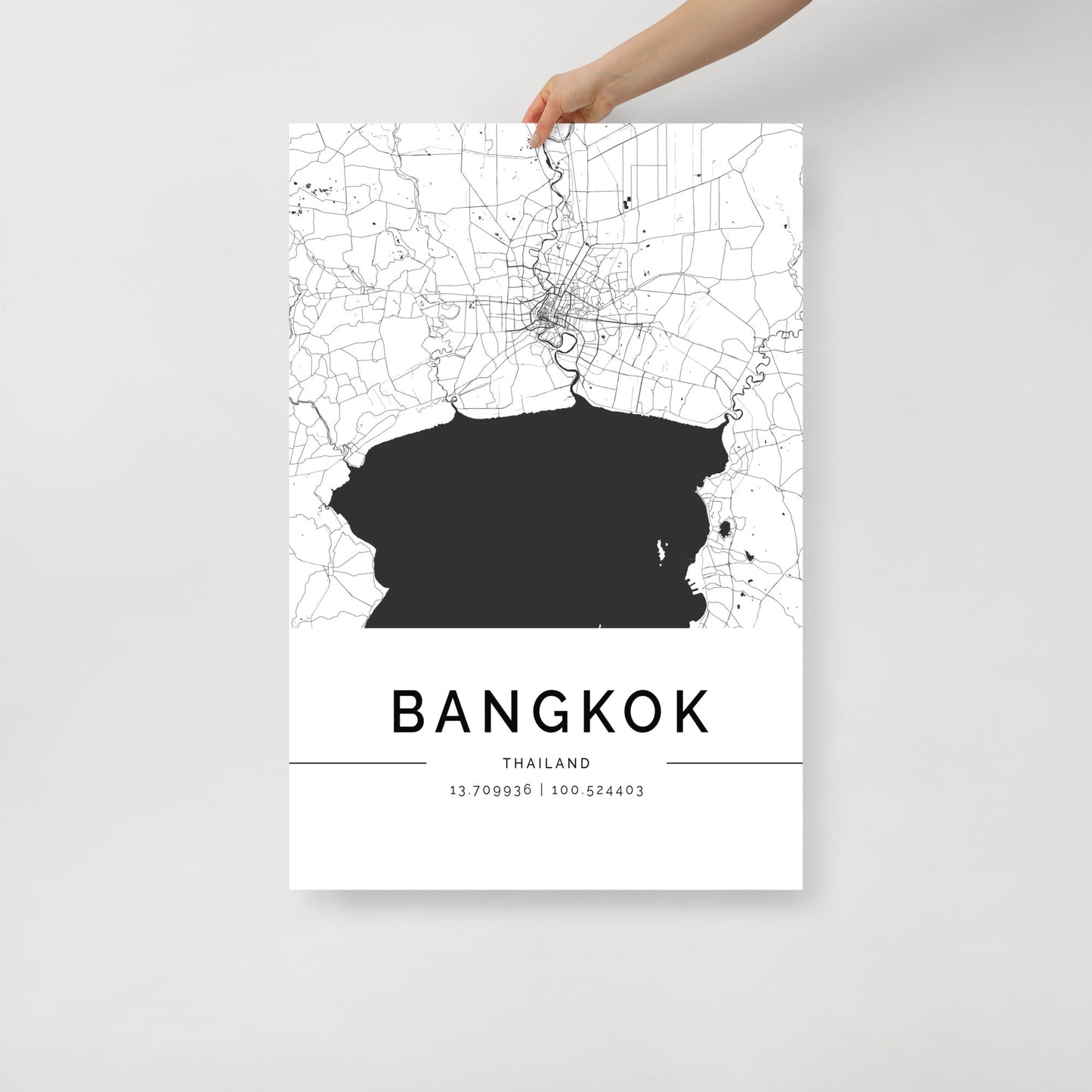 A hand displays a black and white Bangkok Map poster by FN Prints, adorned with geographical coordinates 13.790986 | 100.524403 at the bottom. This striking piece of wall art intricately captures the city's streets and waterways, elegantly showcasing its urban design.