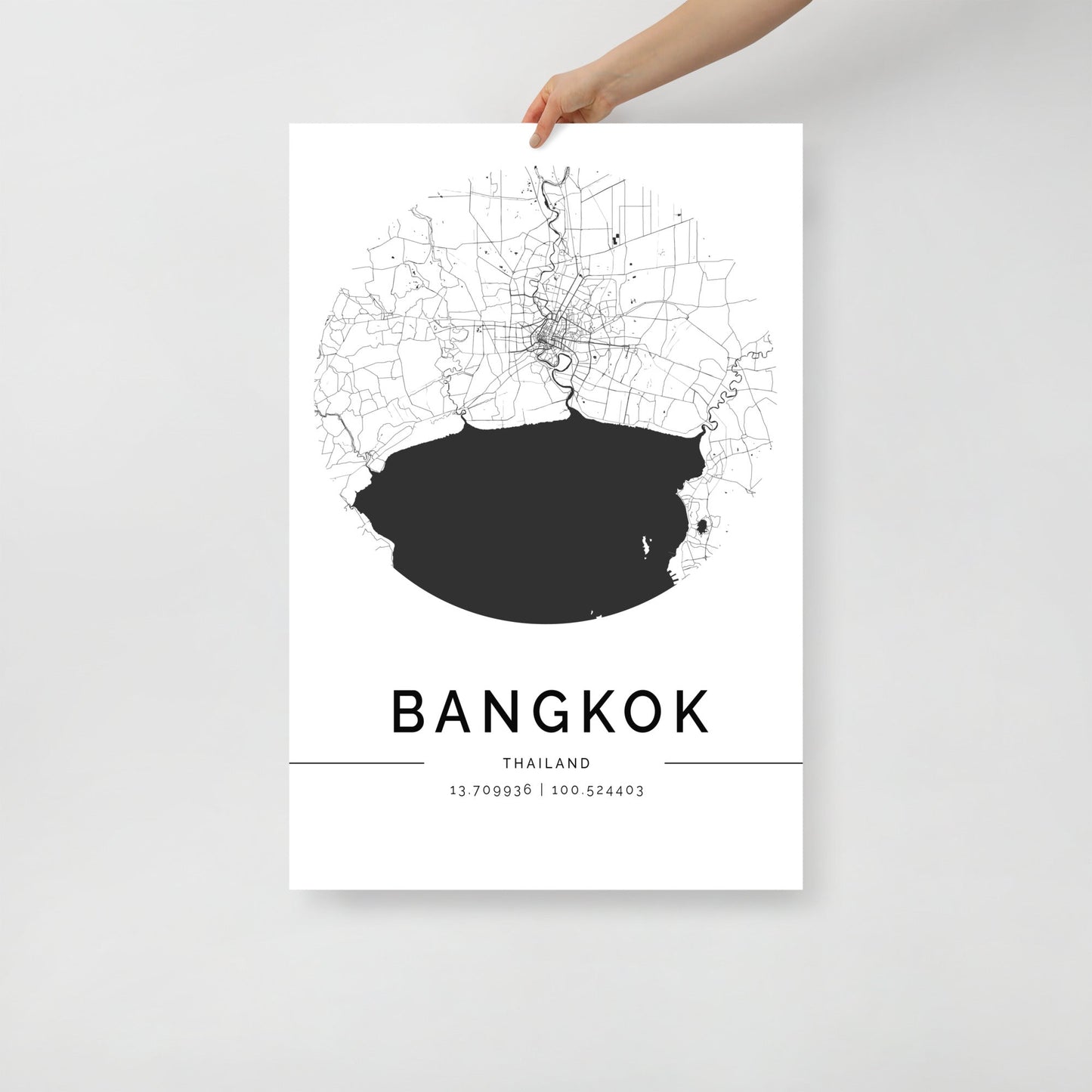 A hand holds a chic Bangkok Map by FN Prints, a piece of urban design wall art featuring minimalist black and white aesthetics. The map showcases city streets with a dark circle indicating the center. Below, "BANGKOK THAILAND" is prominently displayed along with precise map coordinates for added flair.