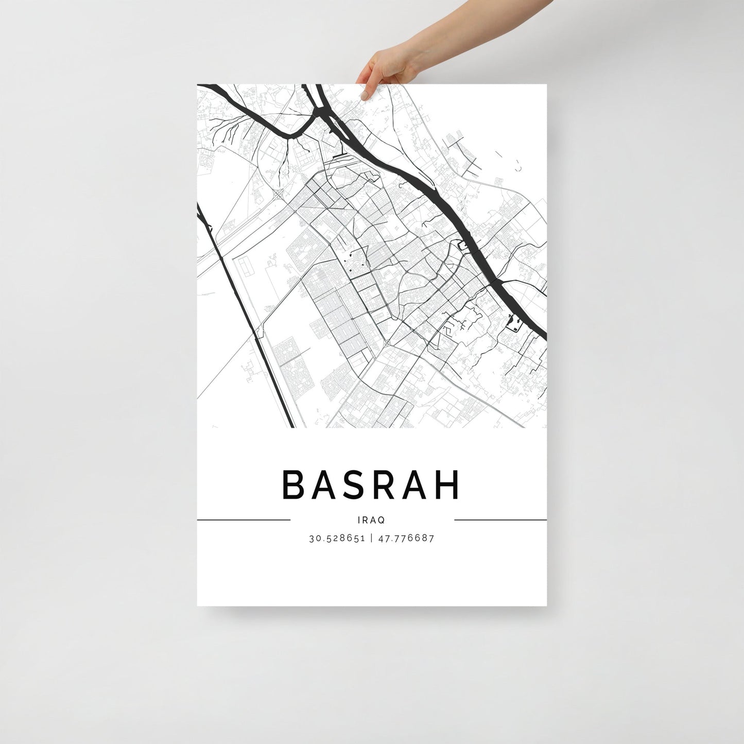 A hand holds the Basra Map by FN Prints, showcasing a monochrome depiction of the city's layout with geographic coordinates 30.528651, 47.776687 below. This minimalist wall art captures the street and road design, offering a glimpse into Middle Eastern history.