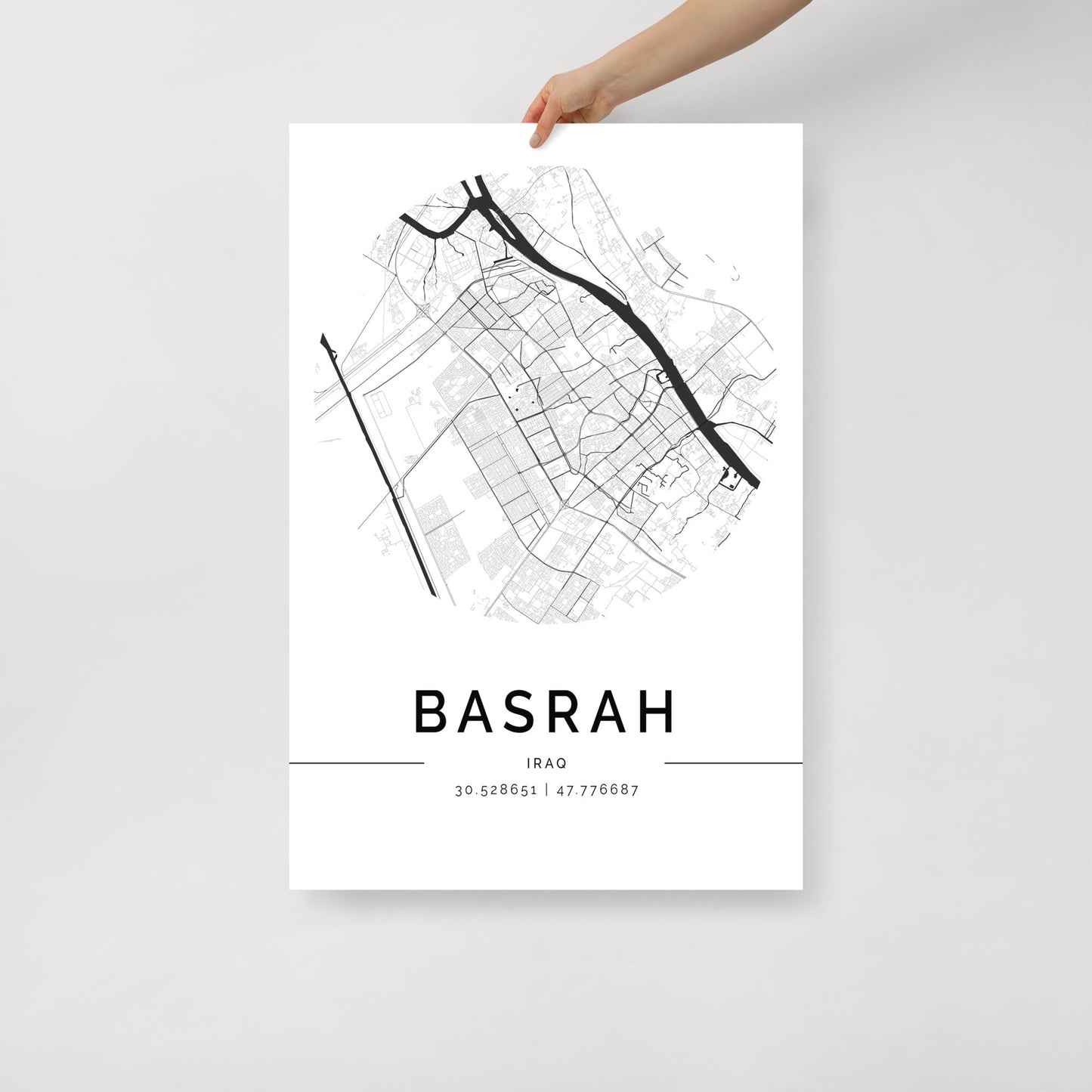 A person holds a minimalist black and white Basra Map art print by FN Prints, showcasing the elegant layout of Basra, Iraq. The streets are detailed with precision and the subtleties of its Middle Eastern history are enhanced by coordinates 30.52865°N, 47.77668°E displayed below the city's name.