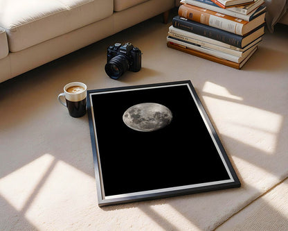 A framed FN Prints photograph titled "Beaver Moon 2024" rests on the carpeted floor beside a cup of coffee and a camera, with several stacked books in close proximity. Gentle sunlight streams through a window, creating celestial-inspired shadows on the carpet.