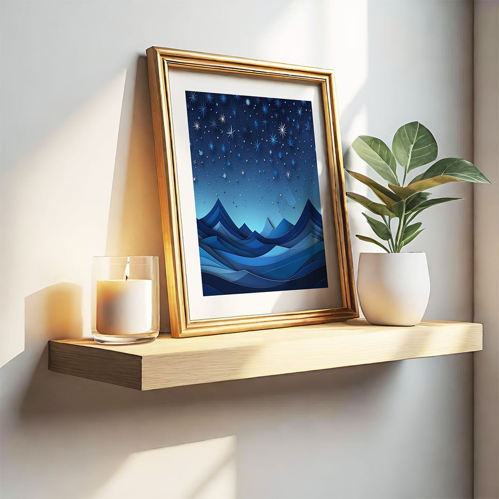 A wooden shelf displays FN Prints' Blues Of Night, a framed artwork depicting a starry night sky over stylized blue mountains, exuding celestial allure. A lit candle in a glass holder and a potted plant sit beside the frame, while sunlight casts shadows on the wall.