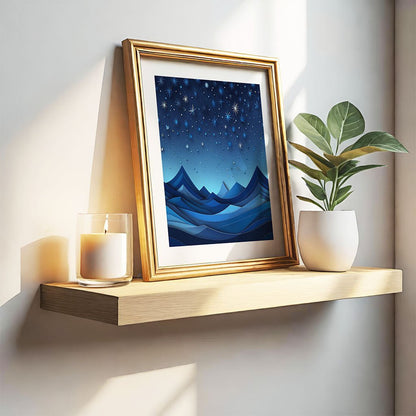 A wooden shelf displays FN Prints' Blues Of Night, a framed artwork depicting a starry night sky over stylized blue mountains, exuding celestial allure. A lit candle in a glass holder and a potted plant sit beside the frame, while sunlight casts shadows on the wall.