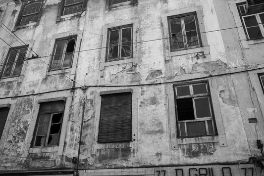 A black-and-white photograph from FN Prints, titled "Broken Windows Black & White," depicting a rundown, multi-story Lisbon building with peeling paint and broken windows. Some windows feature shutters, and there's graffiti adorning the lower part of the wall. The facade is crossed by electrical cables, capturing a sense of vintage urban elegance amid urban decay.