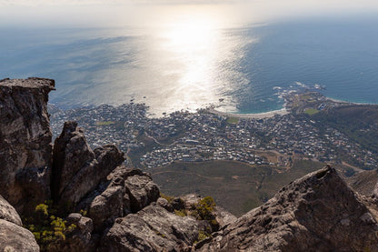 Be captivated by the scenic view captured in "Camps Bay" by FN Prints, showcasing a rocky cliff in Table Mountain National Park. Overlooking Camps Bay with its clustered buildings and houses, the ocean below gleams as sunlight creates a shimmering path along the Western Cape coastline. This print brings to life the stunning mix of green and rocky terrain that characterizes this breathtaking landscape.