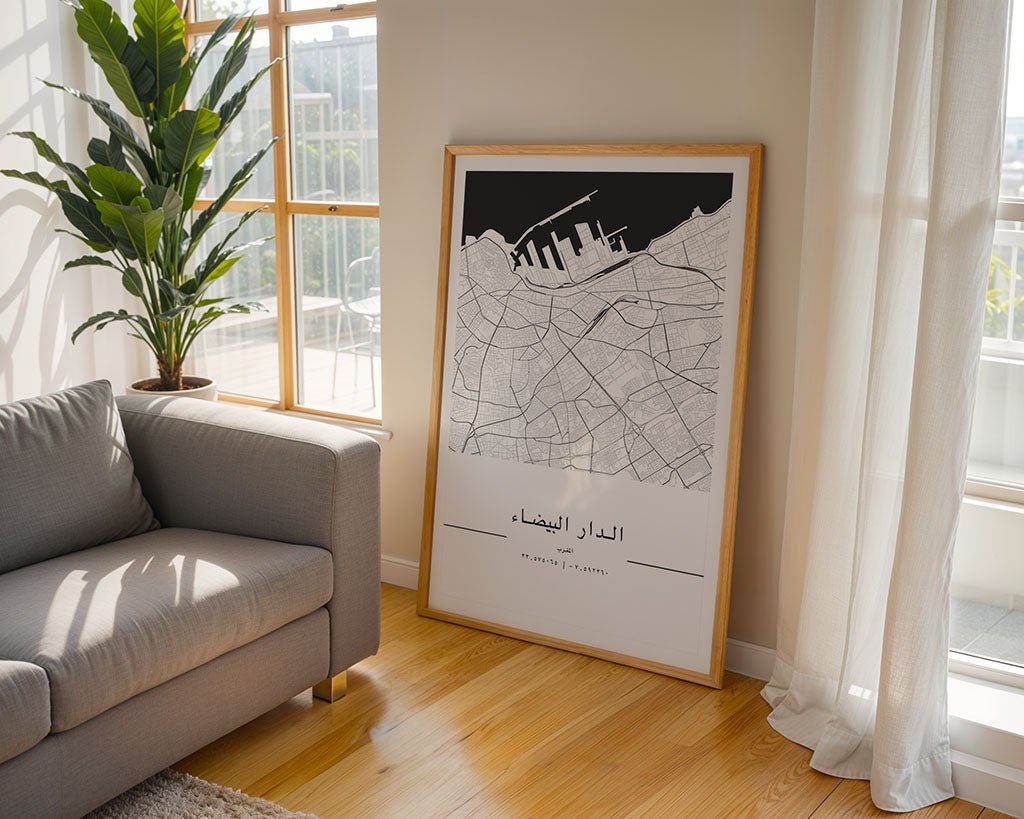 A framed FN Prints Casablanca City Map leans against the wall in a sunlit room, complementing a gray couch and leafy plant. This minimalist print, featuring Arabic script and bathed in light through sheer curtains, makes an ideal housewarming gift for lovers of style and simplicity.