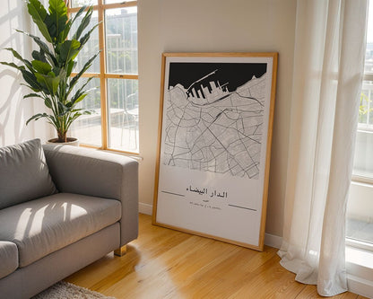 A framed FN Prints Casablanca City Map leans against the wall in a sunlit room, complementing a gray couch and leafy plant. This minimalist print, featuring Arabic script and bathed in light through sheer curtains, makes an ideal housewarming gift for lovers of style and simplicity.