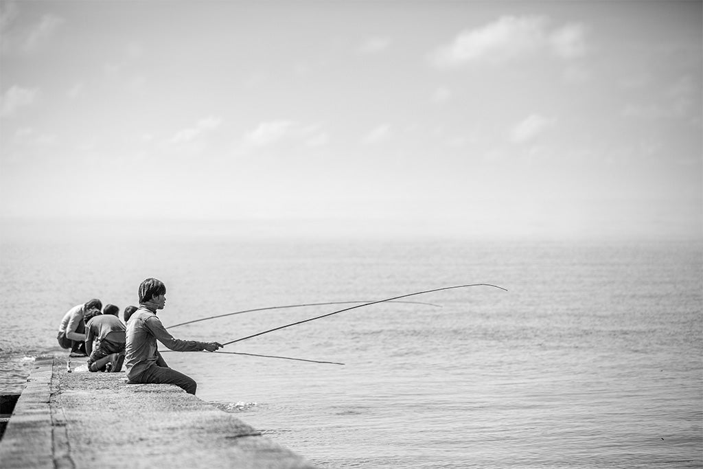 Black and white fine art photography of young Cambodian fishermen sitting on a stone pier with long fishing rods over a calm sea.