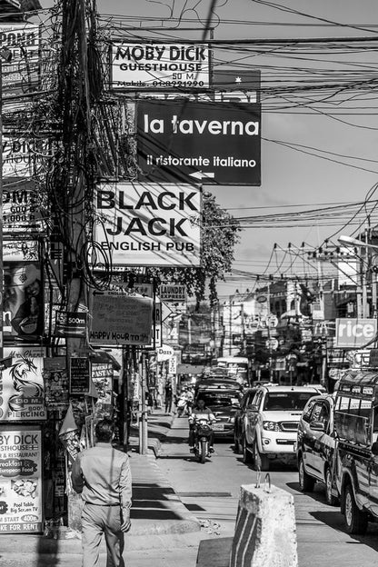 A black and white photograph from FN Prints titled "Chaweng: Koh Samui" captures the bustling urban street of Chaweng, filled with signs for guesthouses, restaurants, and pubs. People walk along the lively street while cars and motorbikes navigate below a maze of overhead wires.