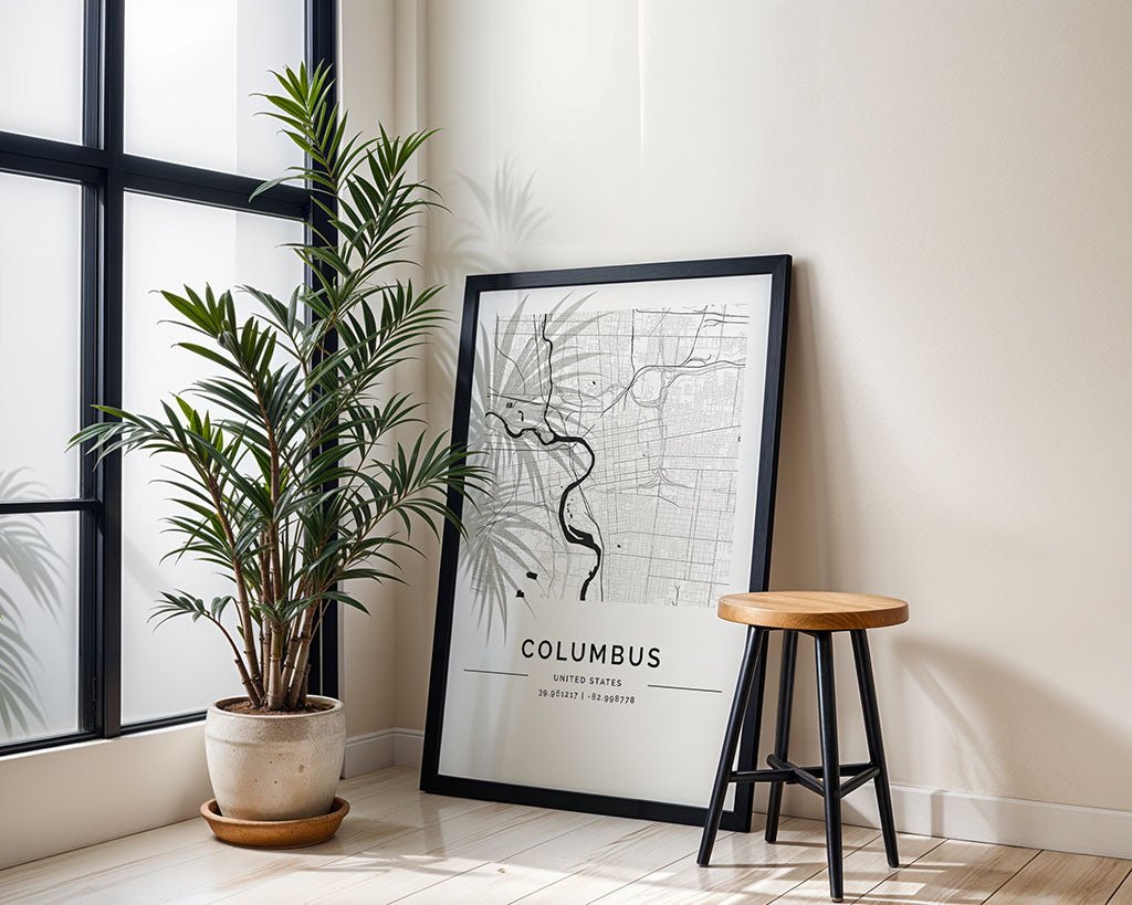 An FN Prints Columbus City Map is propped against the wall beside a shadow-casting potted plant, while a wooden stool with a black base completes the scene in this bright room with large windows.