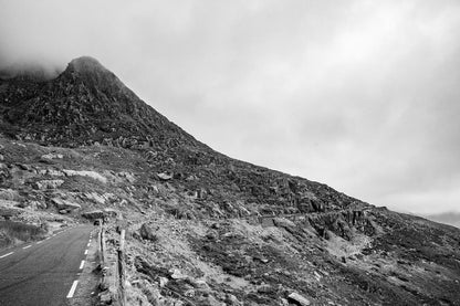 Introducing the FN Prints' Connor Pass Black & White photograph, capturing the majestic mountainous landscape at Connor Pass. This stunning image features a winding scenic route leading up to a rocky peak, partly shrouded in clouds. The rough terrain is dotted with rocks and sparse vegetation, creating a misty and overcast atmosphere that brings this breathtaking scene to life.