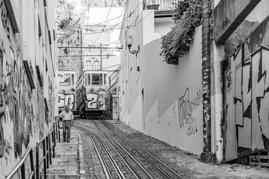 Descending by FN Prints captures an urban exploration scene in black and white, featuring a steep street with two graffiti-covered trams on tram tracks approaching one another. A person traverses down the stairs on the left side, while buildings line the street decorated with lively graffiti artwork.