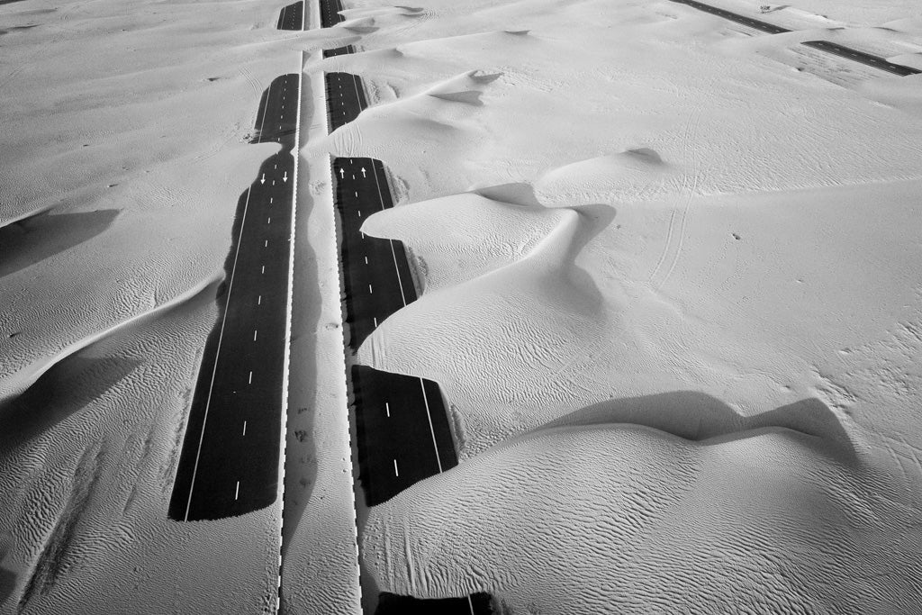 Desert Roads" by FN Prints captures an aerial view of a road flanked by desert sand dunes. The black asphalt contrasts sharply with the light, almost white, sand in this minimalistic elegance. This split highway navigates through the arid desert landscape, with parts of the road partially covered by encroaching sand.