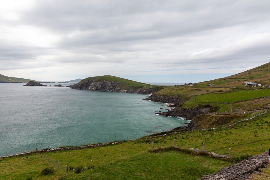 The "Dingle" fine art print by FN Prints captures a scenic coastal landscape with grassy hills sloping towards a rocky shoreline, reminiscent of the Irish coast. The calm blue-green sea meets the land, with small islands visible in the distance and an overcast sky overhead. A few houses dot the hillside, perfectly portraying an authentic Irish coastal scene.