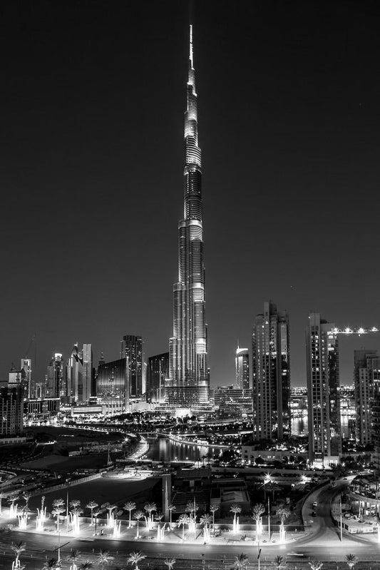 **Dubai Dreams** by **FN Prints** is a captivating black and white fine art photograph showcasing the iconic city skyline at night. A towering, illuminated skyscraper stands prominently amidst several surrounding buildings, with a curved road in the foreground, evoking the breathtaking allure of Dubai's skyline.