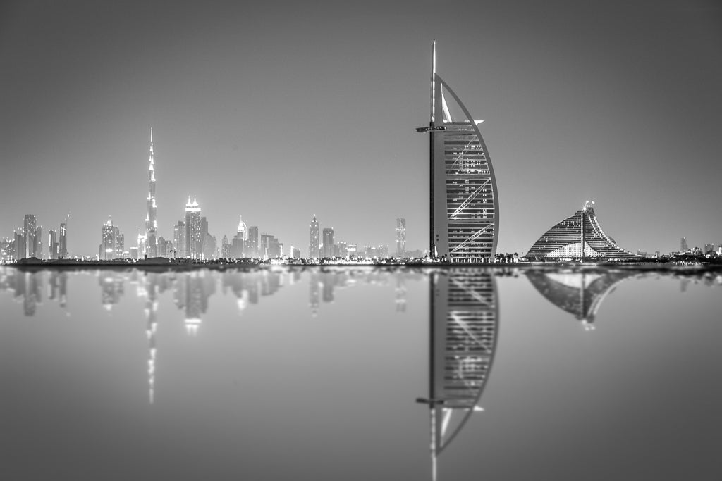 Introducing Dubai Reflections by FN Prints: This black and white cityscape captures the modern skyscrapers of Dubai, including a sail-shaped building, perfectly mirrored on a tranquil body of water. The illuminated skyline against the night sky creates a striking urban scene reminiscent of fine art photography.