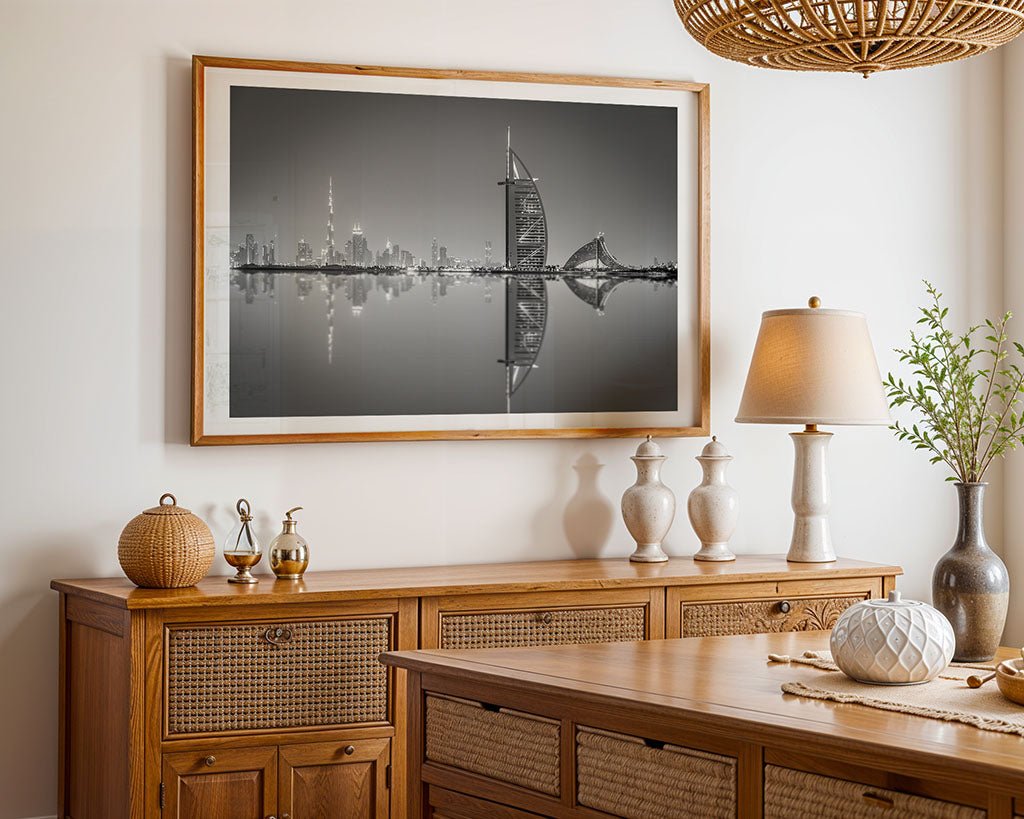 A contemporary dining room is highlighted by "Dubai Reflections," a large framed photograph from FN Prints that showcases the Burj Khalifa in black and white, embodying the elegance of fine art photography. The room also features a wooden sideboard adorned with decorative vases, a table with rattan accents, an elegant lamp, and a lush plant, cultivating a warm, rustic ambiance.