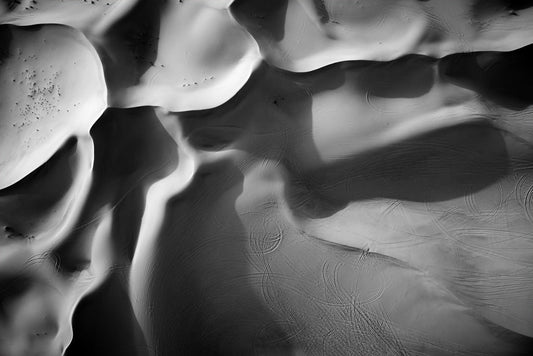 The Dune Part II by FN Prints captures a captivating aerial black and white photograph of a desert landscape, showcasing sand dunes with smooth, flowing patterns. Intricate designs are formed from shadows cast across the surface, complemented by tire tracks that add texture to the scene.